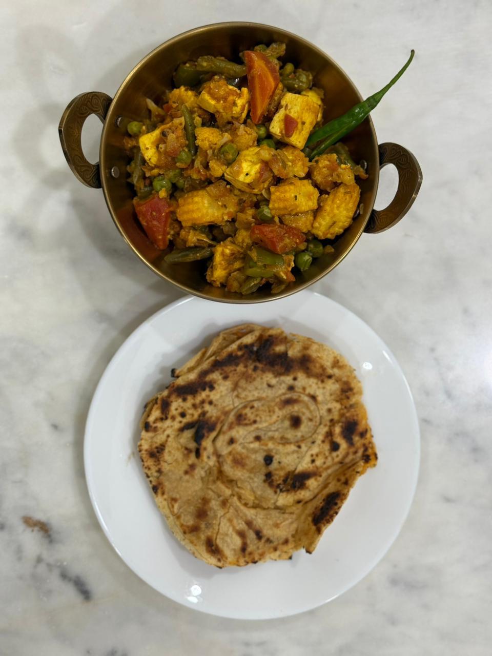 Kadhai of paneer, baby corn, beans and peas beside a laccha paratha on a marble counter