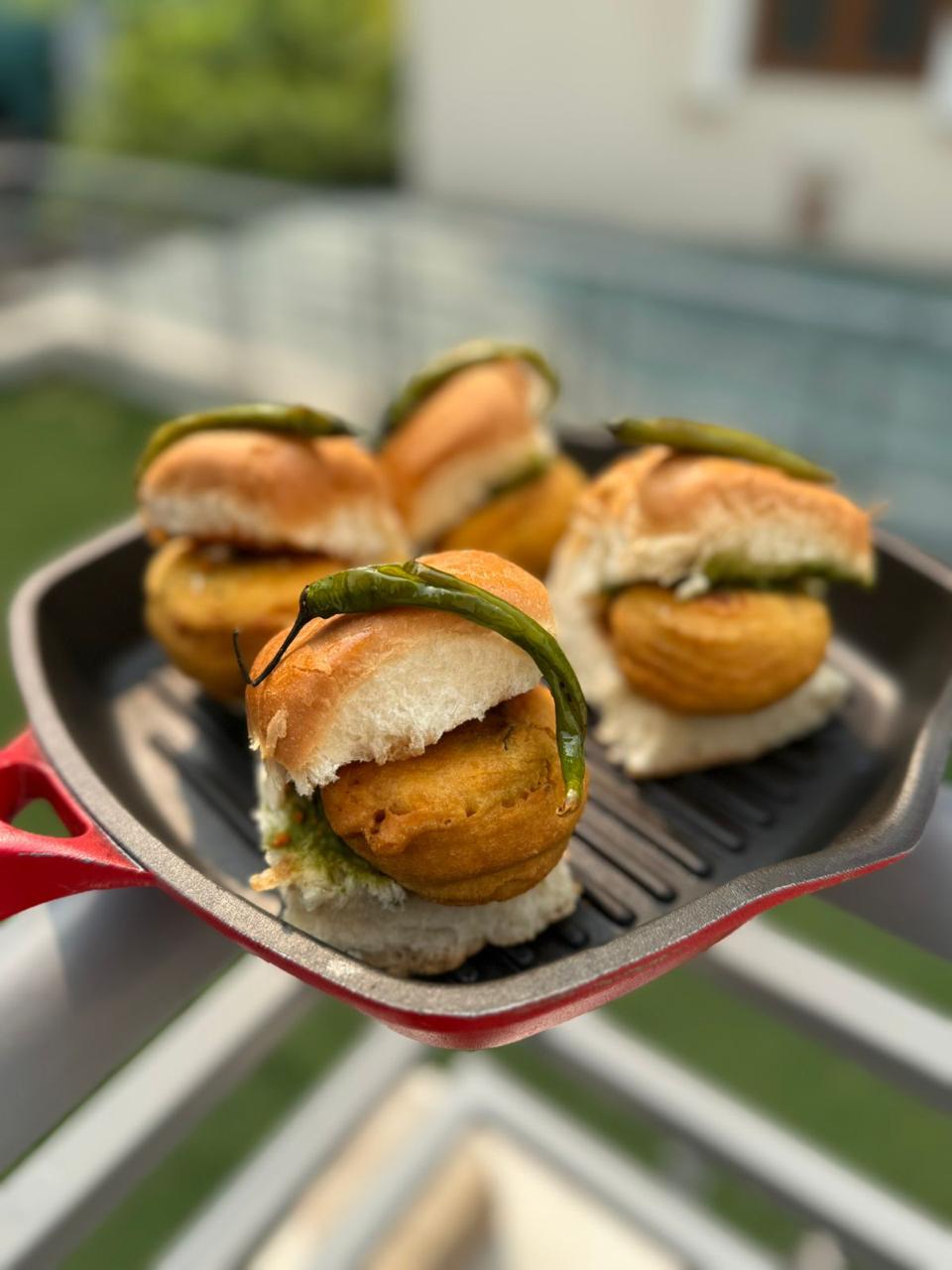 Four vada pao topped with fried green chillies on a cast-iron grill pan