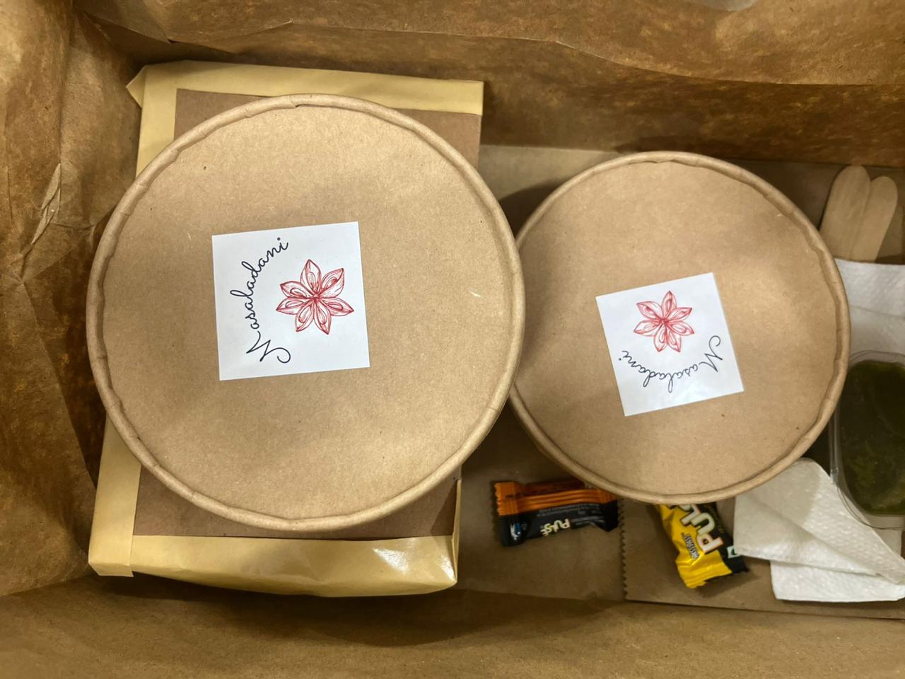 Two kraft bowls sealed with the Masaladani star-anise sticker, packed in a box with cutlery and napkins